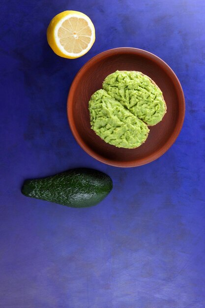 Tostadas de desayuno vegetariano de primer plano con puré de aguacate en un plato de arcilla Tostadas y aguacate verde y limón amarillo sobre un fondo azul Vista desde arriba