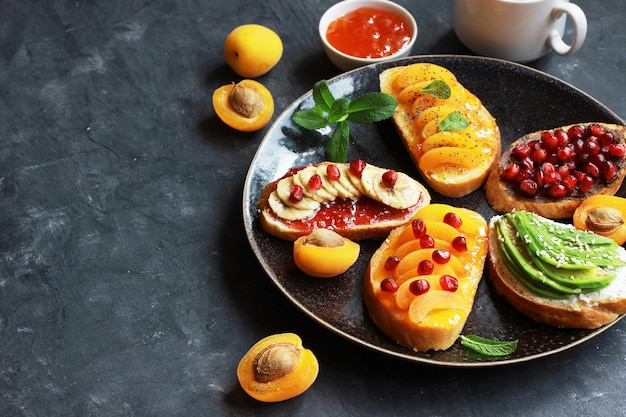 Tostadas dulces con mermelada y varias frutas albaricoques, plátanos, granadas y aguacate en el fondo oscuro copia vista superior del espacio.