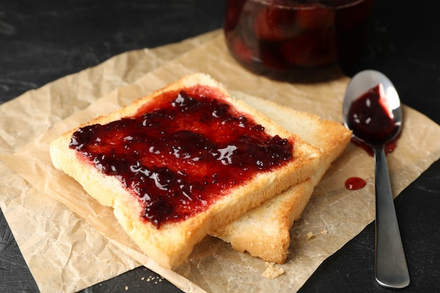 Tostadas y frasco de vidrio con mermelada, cuchara y papel para hornear sobre fondo negro, de cerca