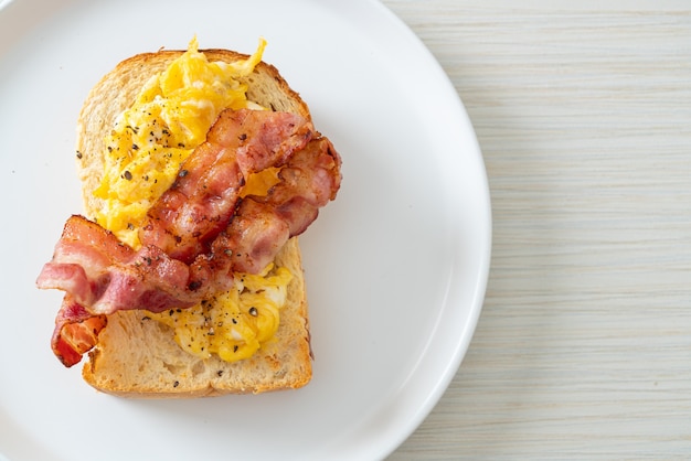 Tostadas de pan con huevos revueltos y tocino en la placa blanca.