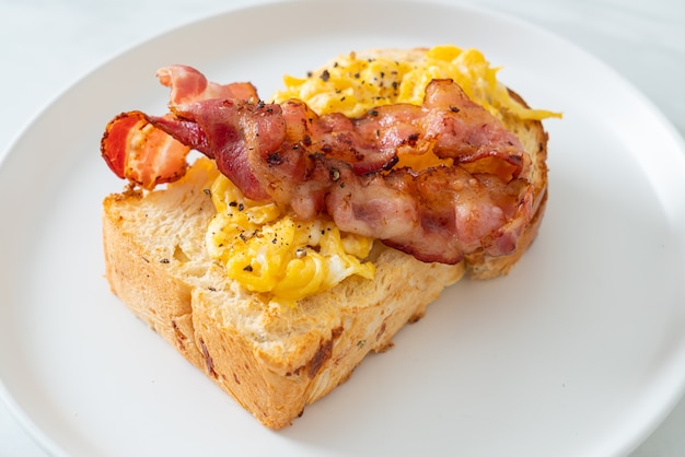 Tostadas de pan con huevos revueltos y tocino en la placa blanca.