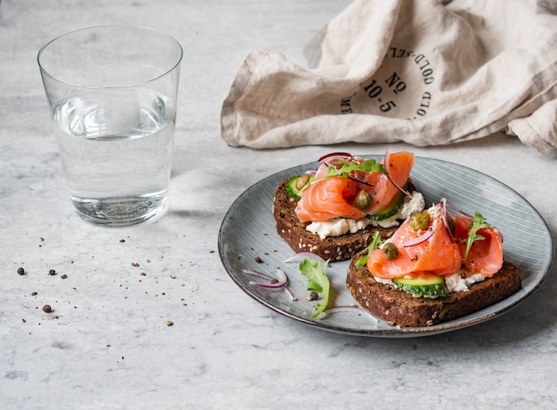 Tostadas saludables con pan de centeno con queso crema, salmón, pepino fresco, alcaparras, semillas de sésamo, pimienta negra y rúcula en un plato. Copia espacio