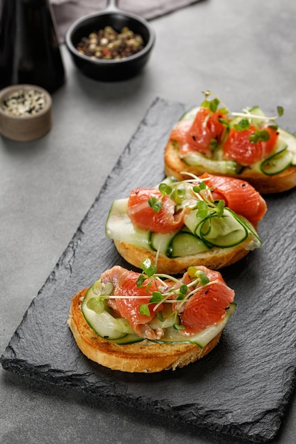 Tostadas saludables con pepinos en rodajas de salmón y microvegetales Aperitivos de bruschetta Sándwiches en tablero de piedra negra y fondo gris