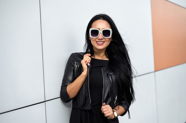 Total schwarzer Fashion Look. Glückliches attraktives Modell mit charmantem Lächeln in der stilvollen Sonnenbrille, die elegantes schwarzes Kleid und modische schwarze Lederjacke mit Fransen trägt.