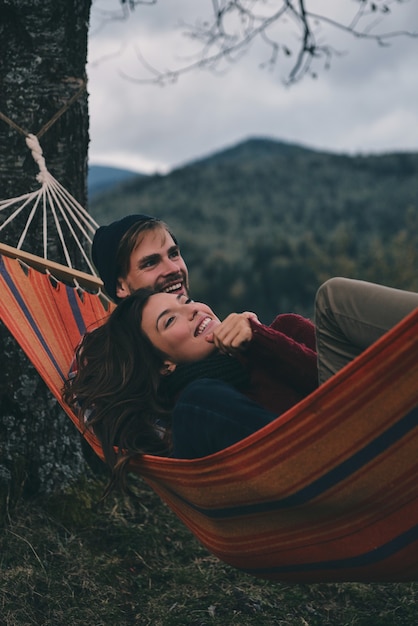 Totalmente feliz. Lindo casal jovem se abraçando e sorrindo enquanto está deitado na rede debaixo da árvore