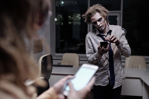 Toter und gruseliger Geschäftsmann mit Zombie-Schmierlack auf Gesicht und Händen, der in der Mitternacht im Smartphone im Büro mit der Frau vorne scrollt