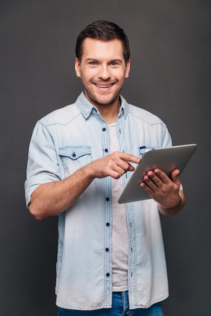 ¡Este touchpad es asombroso! Hombre joven alegre que usa su tableta digital y mirando a la cámara con una sonrisa mientras está de pie contra el fondo gris