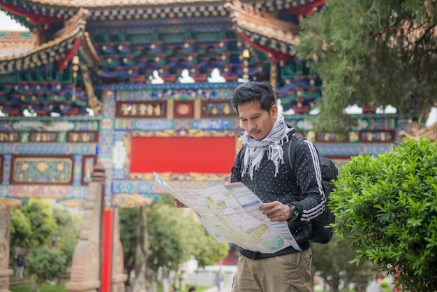 Tourismus des gutaussehenden Mannes, Reisender mit Kamera, Karten, Rucksack und Stativ, chinesischer Architekturhintergrund an Yunnan-Provinz, China