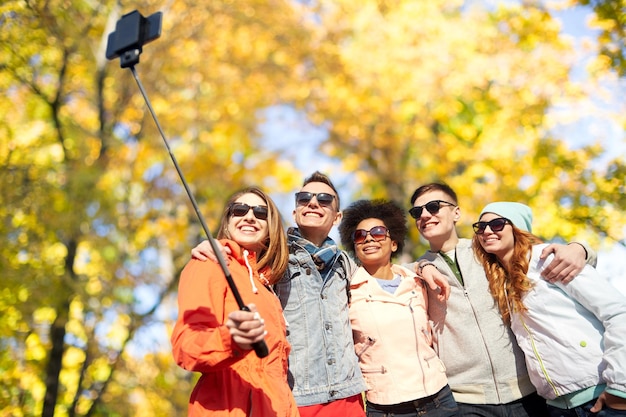 Tourismus-, Reise-, Menschen-, Saison- und Technologiekonzept - eine Gruppe lächelnder Teenager-Freunde, die ein Selfie mit Smartphone und Einbeinstativ über dem Hintergrund des Herbstparks machen