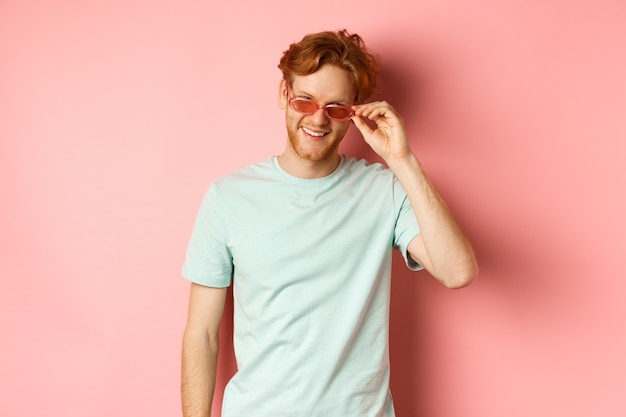 Tourismus- und Urlaubskonzept. Entspannter junger Mann mit roten Haaren, der in Sonnenbrille und T-Shirt steht und erfreut lächelt und über rosa Hintergrund steht.