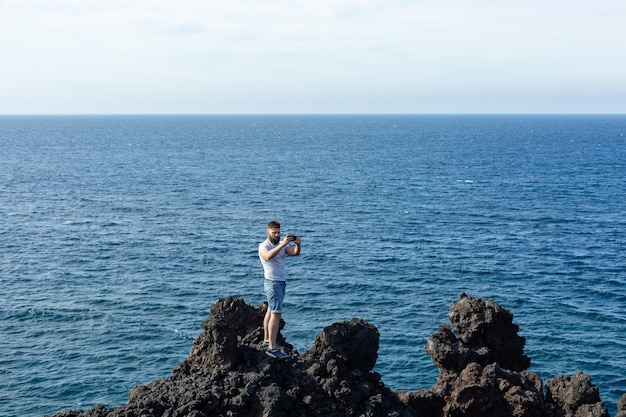 Tourist, der auf einem Felsen auf blauem Hintergrundmeer steht und Foto von seiner Smartphone-Kamera macht. Lanzarote-Insel, Spaine.