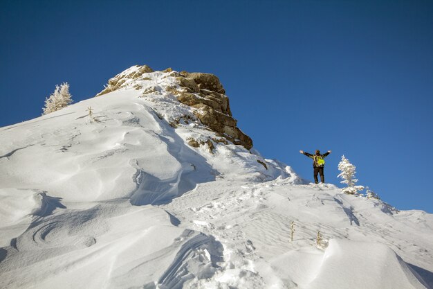 Tourist, der auf schneebedecktem Berggipfel in Siegerhaltung mit erhobenen Händen steht, die Ansicht und Leistung am hellen sonnigen Wintertag genießen.