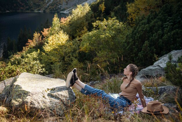 Tourist der jungen Frau, der auf dem Berggipfel und den schönen Bergen sitzt. Landschaft mit sportlichem Mädchen, grünem Wald, Hügeln, Himmel, See. Reisen und Tourismus.