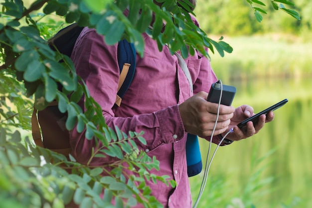 Tourist hält ein Smartphone mit einem tragbaren Ladegerät in den Händen. Mann mit Powerbank lädt das Telefon vor der Kulisse von Natur und Wald auf.