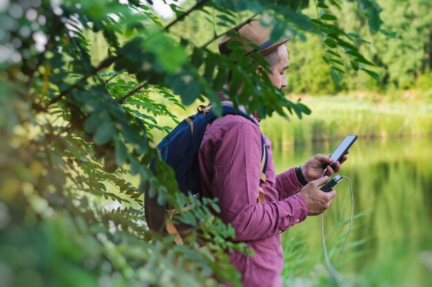 Tourist hält ein Smartphone mit einem tragbaren Ladegerät in den Händen. Mann mit Powerbank lädt das Telefon vor der Kulisse von Natur und Wald auf.