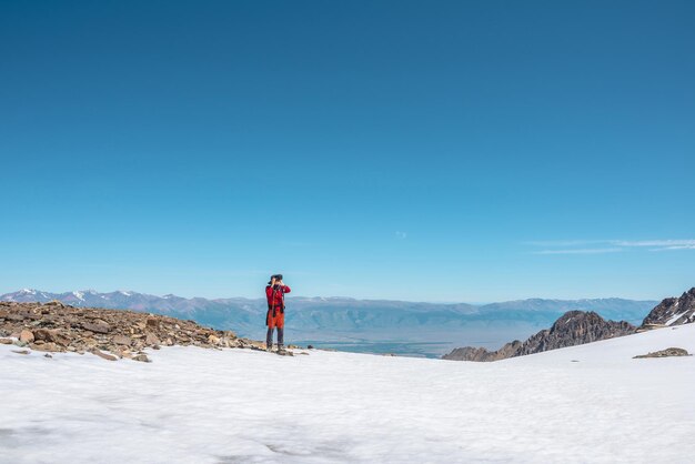 Tourist in Rot geht auf Schneeberg in der Nähe der Abgrundkante in großer Höhe unter blauem Himmel an sonnigen Tagen Mann mit Kamera auf schneebedecktem Berg in der Nähe der Abgrundkante mit Blick auf die große Bergkette in der Ferne