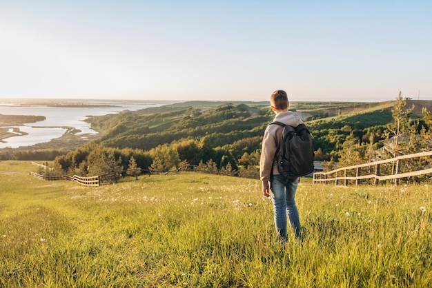 Tourist mit Rucksack, der auf einem Hügel auf der Wiese steht und eine schöne Landschaftsansicht genießt. Rückansicht des Teenager-Wanderers, der in der Natur ruht