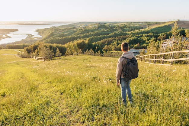 Tourist mit Rucksack, der auf einem Hügel auf der Wiese steht und eine schöne Landschaftsansicht genießt. Rückansicht des Teenager-Wanderers, der in der Natur ruht