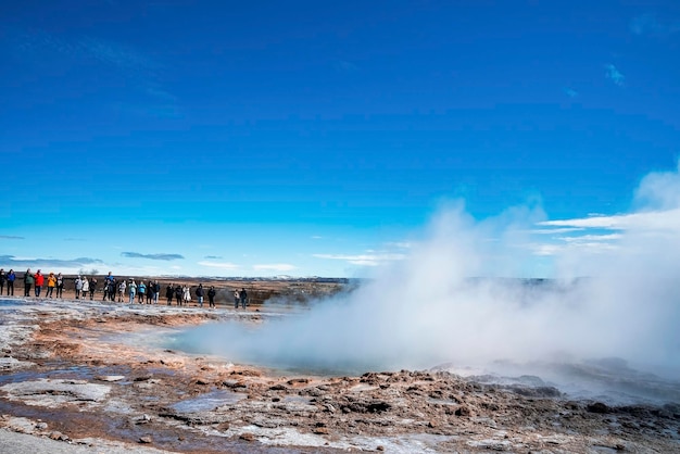 Touristen, die den strokkur-geysirausbruch im tal gegen den blauen himmel erkunden