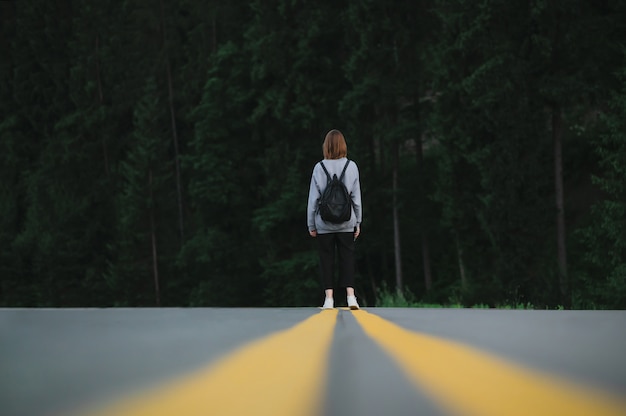 Touristenfrau mit einem Rucksack, der in der Mitte der Autobahn nahe dem Tannenwald steht