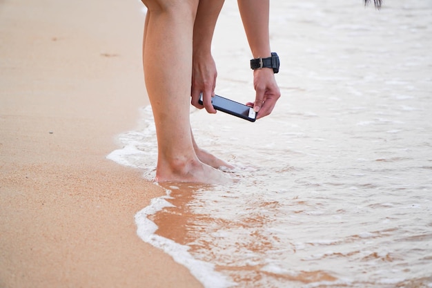 Touristin hält ein modernes Technologie-Smartphone in den Händen und macht ein Selfie-Foto ihrer eigenen Beine, während sie sich im Urlaub an einem Sandstrand niederlegt