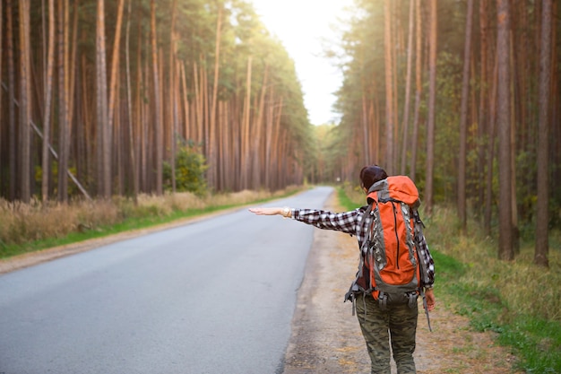 Touristin in einem karierten Hemd mit einem orangefarbenen großen Rucksack in der Nähe einer Autobahn im Wald stimmt für eine Mitfahrgelegenheit. Trampen, Inlandstourismus. Backpacker, Abenteuer allein, Ausflug, Wanderung