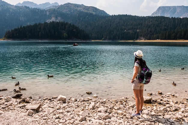 Touristin mit Rucksack und Hut steht am Ufer eines Sees in den Bergen