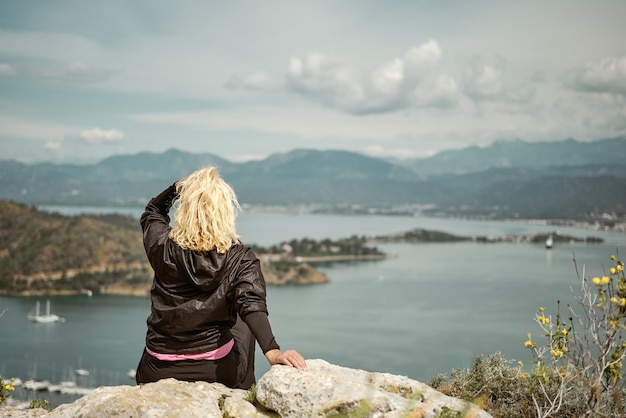 Touristin sitzt auf Felsen in den Frühlingsbergen über der Bucht in der Ägäis, reist allein, wandert und erlebt einen gesunden Lebensstil im Freien, die Natur des Mittelmeers