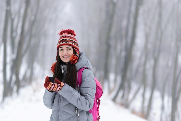 Touristisches brünettes Mädchen mit grauer Jacke, Hut und Fäustlingen, das heißen Tee einer Thermotasse im kalten Winterwald trinkt