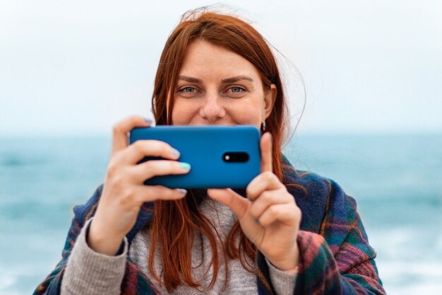 Touristisches kaukasisches Mädchen mit roten Haaren macht Fotos oder Videoaufnahmen auf dem Smartphone am Strand in der Nähe des Ozeans