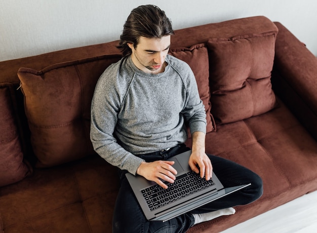 Trabaja desde casa, oficina en casa. Un chico joven con ropa casual está usando una computadora portátil para la comunicación en línea. Se sienta en el sofá, mira la pantalla del portátil y habla.