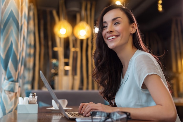 Trabaja en cualquier lugar. Entusiasta enérgica freelancer usando laptop mientras sonríe y mira directamente