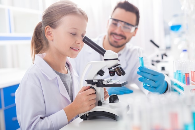 Trabaja con microscopio. Niña hermosa inteligente sonriendo mientras trabaja con microscopio y vestirse en bata de laboratorio