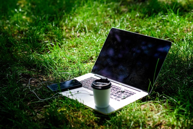 Trabaja y relájate en un entorno natural. Oficina verde. Es la hora del café. Café para llevar. Coffee break al aire libre. Teléfono inteligente moderno portátil con auriculares y taza de café sobre la hierba verde. Parque de verano.