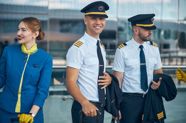 Trabajador de aerolínea joven guapo mirando a cámara y sonriendo mientras espera el vuelo con colegas en el aeropuerto