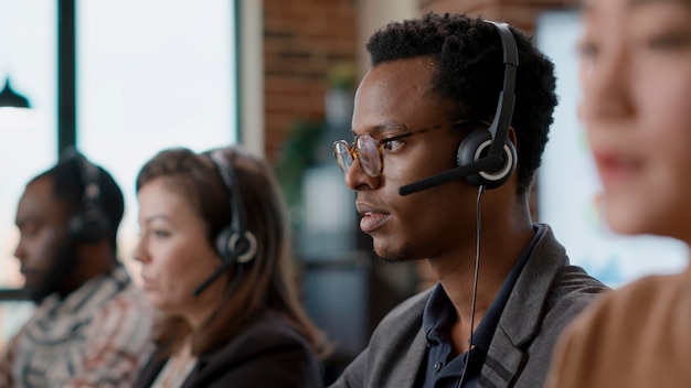 Trabajador afroamericano conversando en auriculares con el cliente, ofreciendo asistencia de telemercadeo en el centro de llamadas. Hombre con auriculares y micrófono trabajando en servicio al cliente. De cerca.