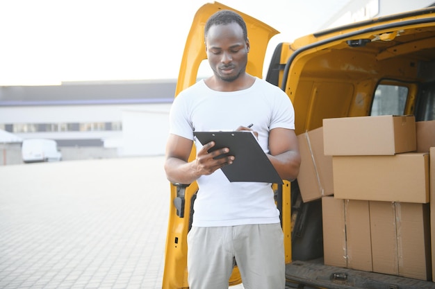 trabajador afroamericano que descarga cajas de la furgoneta al aire libre concepto de servicio de mudanza empleado masculino de una empresa de mudanzas hace un inventario de cajas empacadas y escribe datos en el portapapeles