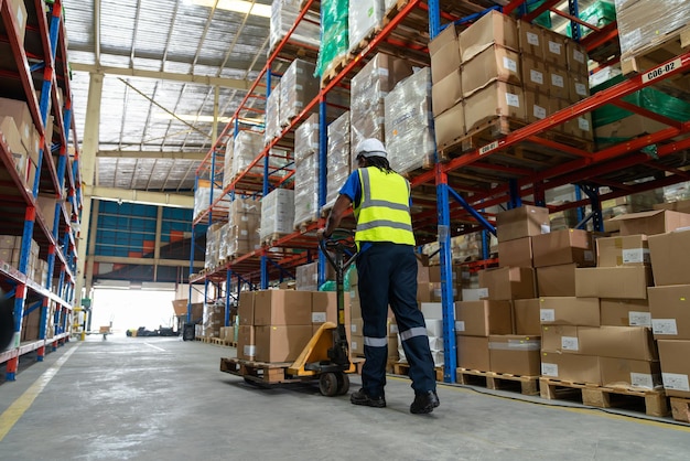 Trabajador afroamericano que usa un camión de paletas para mover cajas de cartón en una fábrica de almacén