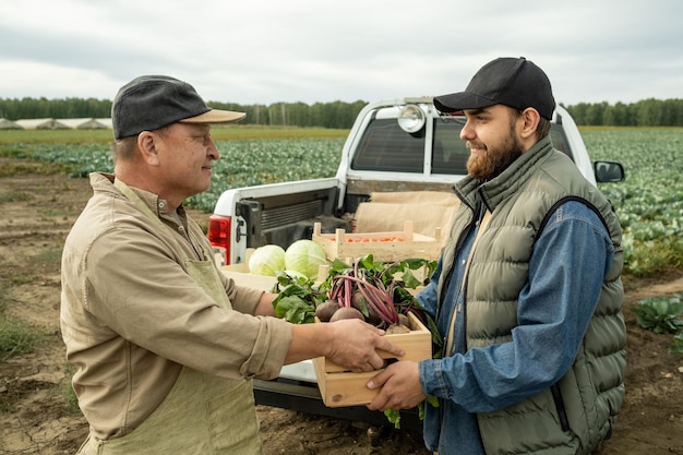 Trabajador agrícola asiático contenido dando caja de verduras frescas al joven conductor de la camioneta pickup