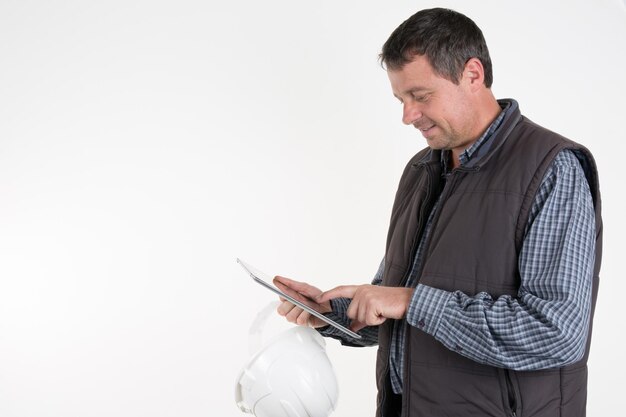 Trabajador alegre con casco blanco y chaleco marrón sosteniendo una tableta digital a prueba de golpes en ambas manos.