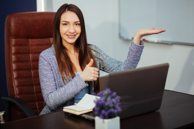 Trabajador alegre escribiendo en la computadora portátil y mirando a otro lado con una sonrisa brillante. Ella está sentada en un escritorio en una oficina. Retrato