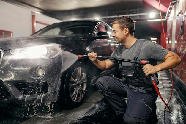 Un trabajador alegre del lavado de coches está usando una pistola de agua y lavando un coche sucio en la estación