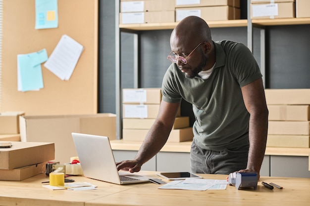 Trabajador de almacén africano escribiendo números de pista de paquetes en la computadora portátil en la mesa en la sala de almacenamiento