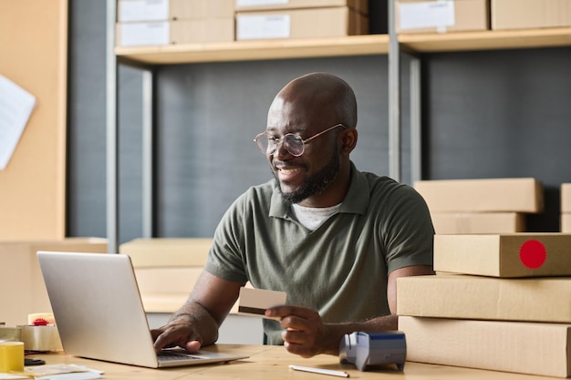 Trabajador de almacén africano pagando en línea con tarjeta de crédito escribiendo en la computadora portátil en la mesa