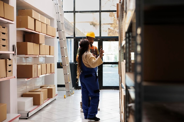 Trabajador de almacén afroamericano escaneando mercancías en stock con el gerente. Todo el equipo de la sala de almacenamiento negro realiza un inventario de productos de la tienda minorista utilizando un escáner de código de barras y una tableta digital