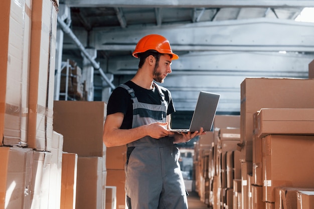 Trabajador de almacenamiento en portátil uniforme y moderno en manos comprueba la producción.