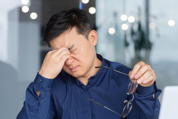 Trabajador asiático cansado en el trabajo tiene dolor en los ojos hombre de negocios con anteojos trabajando hasta tarde usando una computadora portátil en el trabajo