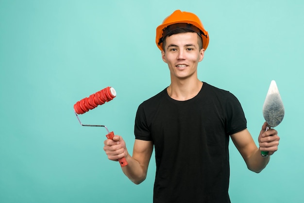 Un trabajador asiático con un casco naranja se para con un rodillo de pintura y una paleta en una imagen de fondo azul