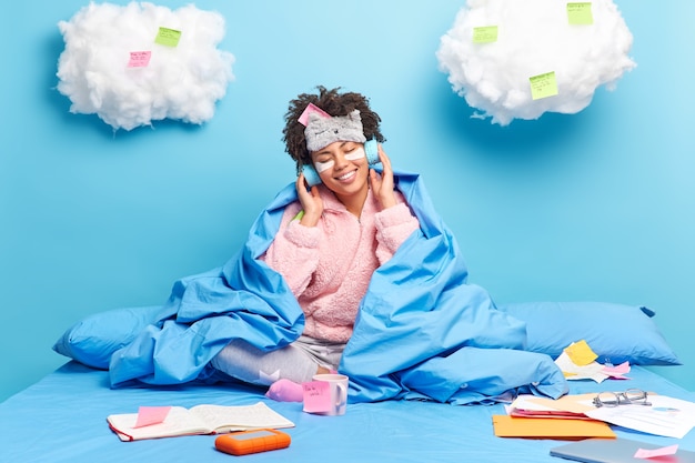 Foto trabajador autónomo trabaja desde casa se sienta en la cama viste pijama escucha música con auriculares hace notas utiliza pegatinas aisladas en azul