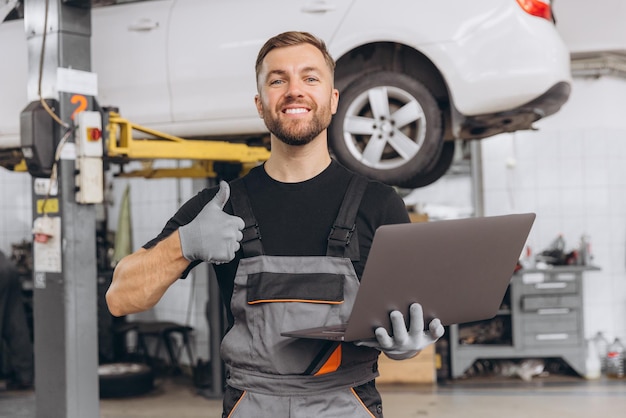 Trabajador de autoservicio hombre barbudo sonriente sosteniendo la computadora portátil en las manos y mostrando los pulgares hacia arriba mecánico reparando automóvil levantado detrás